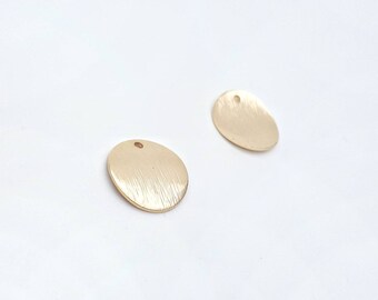 May include: Two gold-colored, oval-shaped metal pendants with a brushed texture and a small hole at the top for stringing. The pendants are set against a white background.