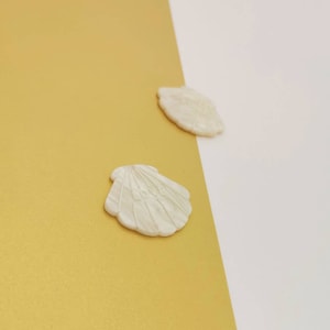 Op de afbeelding: Twee witte schelpvormige decoraties. Op een schelp staat het woord "LOVE" gegraveerd. De schelpen staan tegen een gouden en witte achtergrond. De schelpen zijn waarschijnlijk gemaakt van keramiek of een vergelijkbaar materiaal.