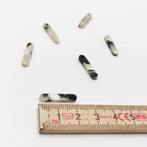 K&ouml;nnte beinhalten: F&uuml;nf rechteckige Anh&auml;nger aus marmoriertem schwarzem und wei&szlig;em Kunststoff mit einem Loch oben. Die Anh&auml;nger sind etwa 3,8 cm lang. Ein Lineal ist im Bild als Gr&ouml;&szlig;enreferenz zu sehen.