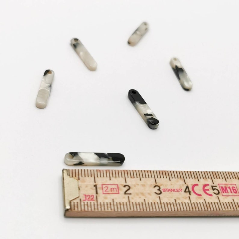 K&ouml;nnte beinhalten: F&uuml;nf rechteckige Anh&auml;nger aus marmoriertem schwarzem und wei&szlig;em Kunststoff mit einem Loch oben. Die Anh&auml;nger sind etwa 3,8 cm lang. Ein Lineal ist im Bild als Gr&ouml;&szlig;enreferenz zu sehen.