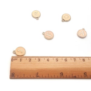 May include: Five gold-colored metal charms with a light pink enamel finish. Each charm features a different letter of the alphabet: A, B, C, E, and J. The charms are arranged on a white surface with a wooden ruler for scale.