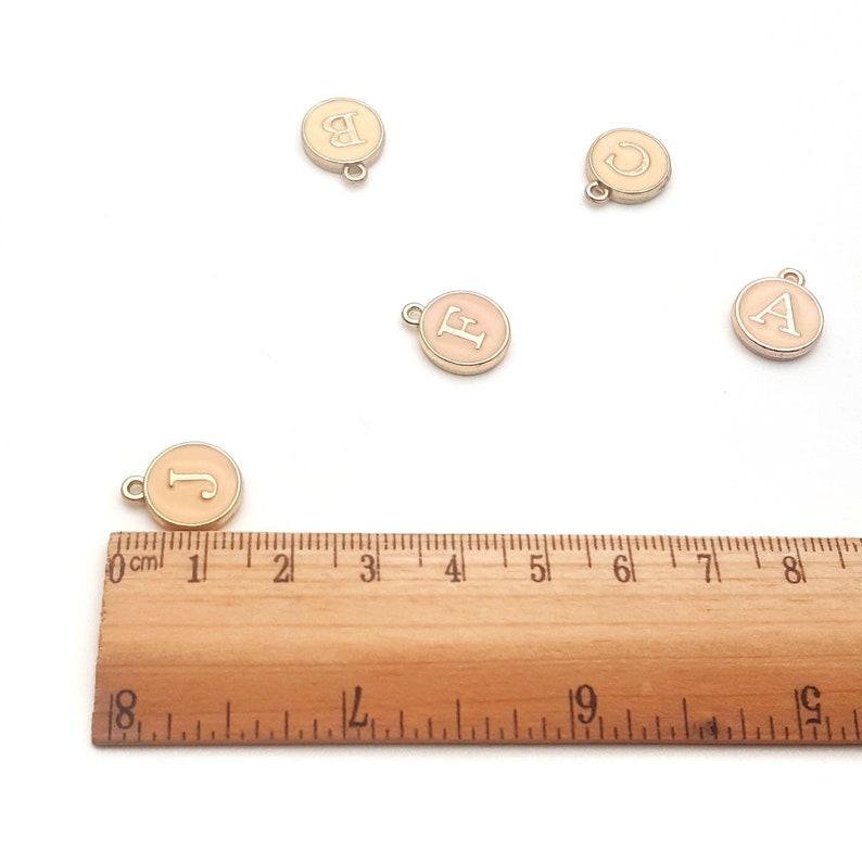 May include: Five gold-colored metal charms with a light pink enamel finish. Each charm features a different letter of the alphabet: A, B, C, E, and J. The charms are arranged on a white surface with a wooden ruler for scale.