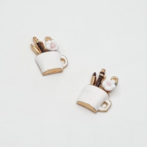 May include: Two white enamel charms shaped like mugs, each with gold trim and handles. The mugs hold brown pencils and a small white flower with a pink centre. These charms are ideal for jewellery making or craft projects.