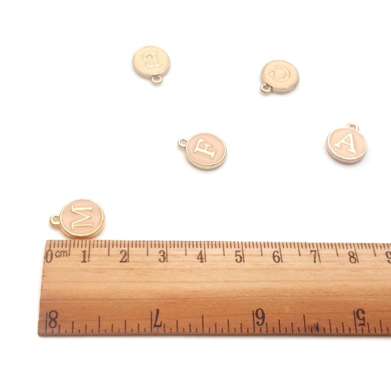 May include: Round, gold-colored charms with letters A, B, C, F, and M in a light pink color. Each charm has a small loop at the top. A wooden ruler is placed near the charms, with measurements in inches.