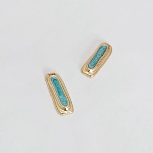 May include: A pair of gold-coloured rectangular earrings, each featuring a light blue marbled enamel inlay. The earrings have a simple, elegant design. The earrings are set against a white background.