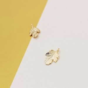 May include: Two gold-coloured leaf charms, one larger than the other, are displayed on a white and yellow background.
