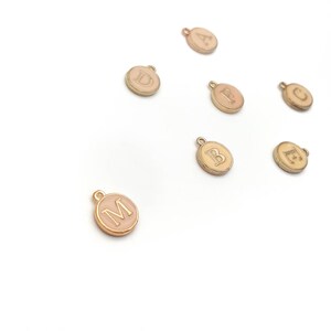 May include: A collection of gold-colored, round charms with a light pink enamel center. Each charm features a different letter of the alphabet in a serif font. The charms are scattered on a white background, with a small loop at the top for attaching to jewelry.