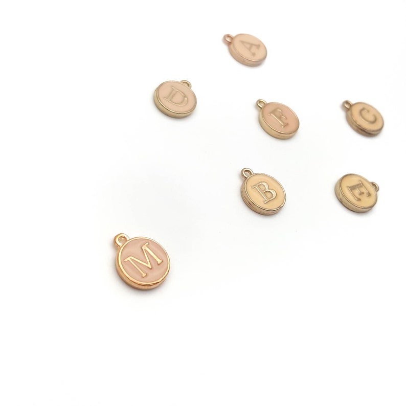May include: A collection of gold-colored, round charms with a light pink enamel center. Each charm features a different letter of the alphabet in a serif font. The charms are scattered on a white background, with a small loop at the top for attaching to jewelry.