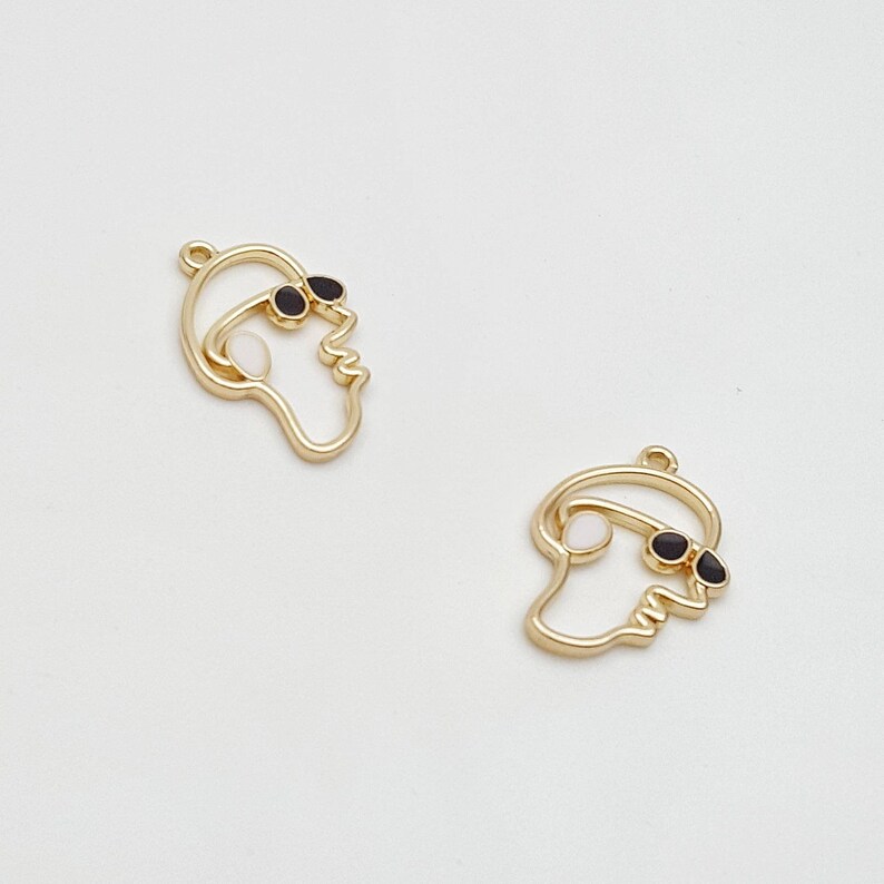image shows two outline face charms in shiny gold tone metal, the face has a sunglasses detail and a hoop for attachment