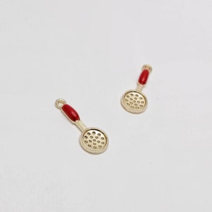 May include: Two gold-toned metal charms shaped like kitchen skimmers or slotted spoons. Each charm features a circular head with a perforated grid pattern and a handle accented with a smooth, solid red enamel inlay. A small loop is at the top.