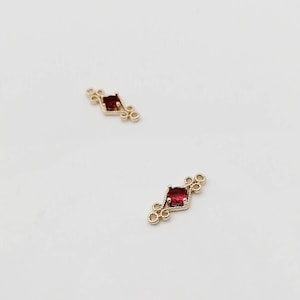 Puede incluir: Dos conectores de metal dorado con una piedra preciosa roja en el centro de cada uno. Los conectores tienen forma de diamante con pequeños círculos a cada lado.