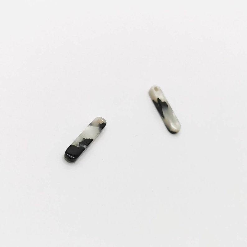 K&ouml;nnte beinhalten: Zwei rechteckige Charms aus marmoriertem schwarzem und wei&szlig;em Kunststoff mit einem Loch oben.
