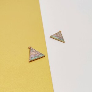 Puede incluir: Dos colgantes triangulares dorados con diseños de esmalte pastel. Cada colgante presenta un motivo de luna creciente y nubes en tonos rosa, morado y turquesa. Los colgantes están sobre un fondo dorado y blanco.