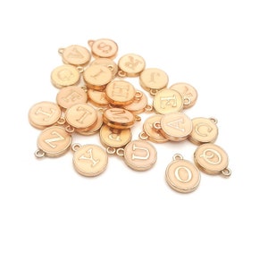 May include: A collection of gold-toned metal charms with letters of the alphabet printed on them. The charms are round and have a light pink enamel finish.