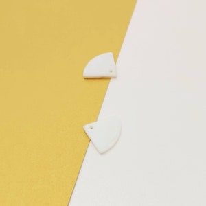 May include: Two white, semi-circular pendants with a small hole at the top, set against a gold and white background. The pendants have a smooth, glossy finish and a curved edge.