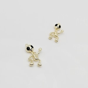 Pode incluir: Dois pingentes de esqueleto de ouro com olhos de esmalte preto. Os esqueletos estão em uma pose de dança com os braços levantados.