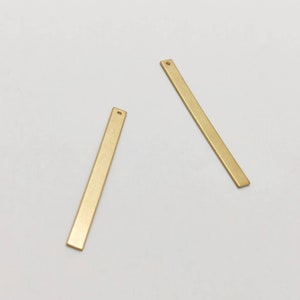 Könnte beinhalten: Zwei goldfarbene, rechteckige Metallstäbe mit einem kleinen Loch an einem Ende, die auf einer weißen Oberfläche liegen. Die Stäbe sind dünn und haben eine matte Oberfläche, geeignet für die Schmuckherstellung oder das Basteln.