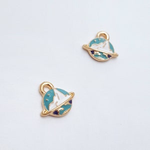 May include: Two gold-coloured planet charms with a light blue and white design. Each charm has a small loop at the top for attaching to jewellery. The planet has a gold ring around it and a dark blue accent.