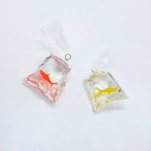 May include: Two clear plastic bags with gold fish charms hanging from silver jump rings. The fish in the left bag is orange and the fish in the right bag is yellow.