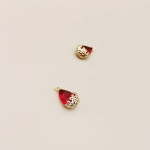 May include: Two red glass teardrop-shaped charms with gold floral accents. The charms have a loop at the top for attaching to jewellery.