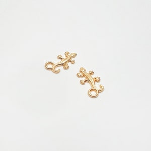 Op de afbeelding: Twee goudkleurige metalen hagedisbedels. Elke bedel heeft een gedetailleerd hagedisontwerp met een gekrulde staart en kleine pootjes. De bedels staan op een witte achtergrond.