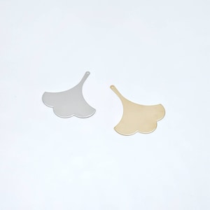 Peut inclure: Deux pendentifs en métal en forme de feuille. L'un est argenté, l'autre doré. Les deux ont un bord festonné et un petit trou pour l'enfilage. Les pendentifs sont sur fond blanc.