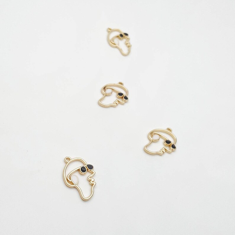 May include: Four gold-colored metal charms in the shape of a face wearing sunglasses. The sunglasses are black. The charms are arranged on a white background. These charms are suitable for jewelry making.
