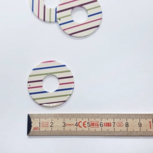 May include: Two white ceramic circles with a hole in the center. The circles are decorated with stripes of blue, green, pink, and purple. The circles are about 2 inches in diameter.