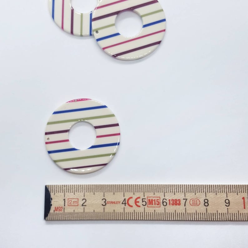 May include: Two white ceramic circles with a hole in the center. The circles are decorated with stripes of blue, green, pink, and purple. The circles are about 2 inches in diameter.