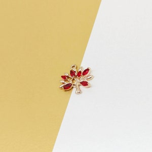 Può includere: Un ciondolo a forma di albero color oro con foglie di gemme rosse. Il ciondolo ha un piccolo anello nella parte superiore per il fissaggio a una collana o un braccialetto. Lo sfondo è diviso diagonalmente, con una combinazione di colori oro e bianco.