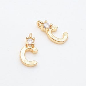 May include: Two gold-coloured letter "C" charms, each featuring a small, clear gemstone at the top. These charms have a loop for attaching to jewellery or crafts. The background is plain white.