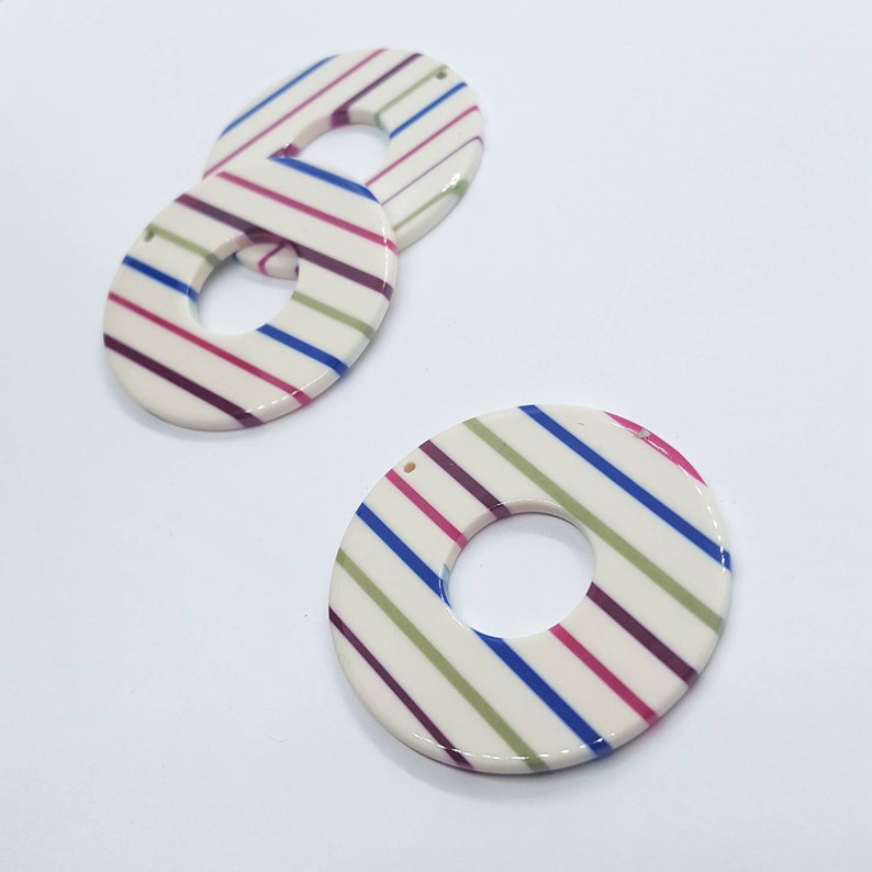 May include: Three white, round, striped, acrylic, decorative pieces with a hole in the center. The stripes are green, blue, pink, and purple.