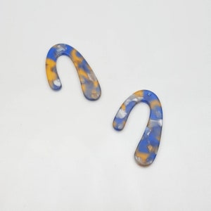 Op de afbeelding: Twee abstracte, gebogen oorbellen in een gemarmerd patroon van blauw, geel en wit. De oorbellen hebben een gladde, glanzende afwerking en een klein gaatje aan de bovenkant voor bevestiging.