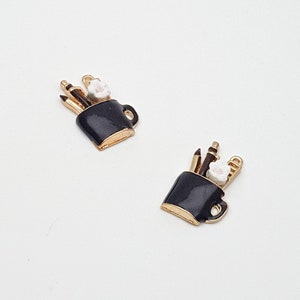 May include: Two black enamel charms shaped like mugs, each with gold trim and handles. The mugs contain pencils, a paintbrush, and a small white flower. These charms are suitable for jewellery making or craft projects.