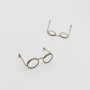 Może przedstawiać: Dwie pary małych, okrągłych okularów z cienkimi, złotymi drucianymi oprawkami. Soczewki są przezroczyste. Jedna para jest ostra, a druga lekko rozmyta w tle. Okulary są umieszczone na białym tle.