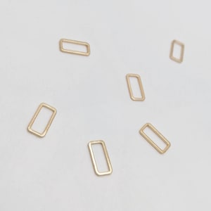 May include: Six gold-coloured metal jump rings, all rectangular in shape, are arranged on a white surface.