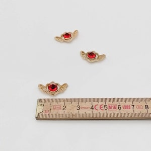 May include: Three gold-colored charms with red rhinestone centers. The charms are shaped like wings and are approximately 1 cm in length. A ruler is in the background for size reference.