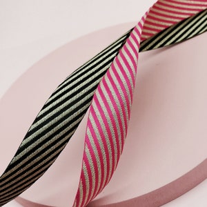 Könnte beinhalten: Zwei gestreifte Bänder, eines schwarz und gold, das andere pink und gold, sind auf einer rosa Oberfläche übereinander gelegt.