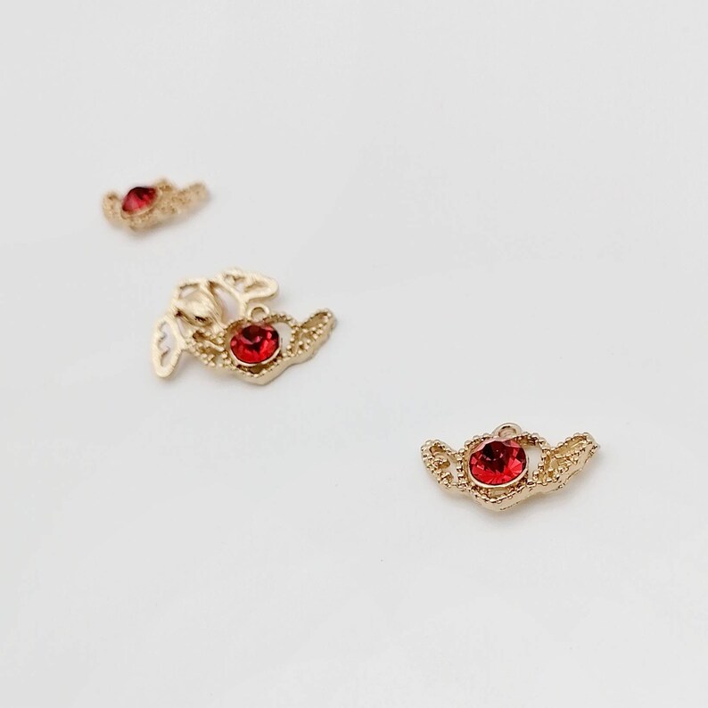 May include: Three gold-tone metal charms with red rhinestone accents. Each charm features a delicate, ornate design.
