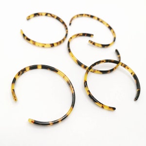 May include: Several open-ended bangles with a tortoiseshell pattern are scattered on a white surface. The bangles are a mix of black, brown, and yellow, with a glossy finish. The open design allows for easy wear.