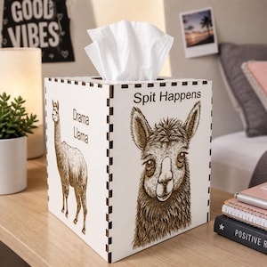May include: A white wooden tissue box with a llama design and the phrases "Drama Llama" and "Spit Happens" etched on the sides. The box has a square shape with a decorative edge and a slot for tissue dispensing.