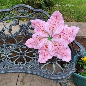 May include: A pink crocheted lily flower cushion resting on a black metal garden bench. The bench is on a stone patio. The text "Pippa Patterns" is visible in the upper right corner of the image.