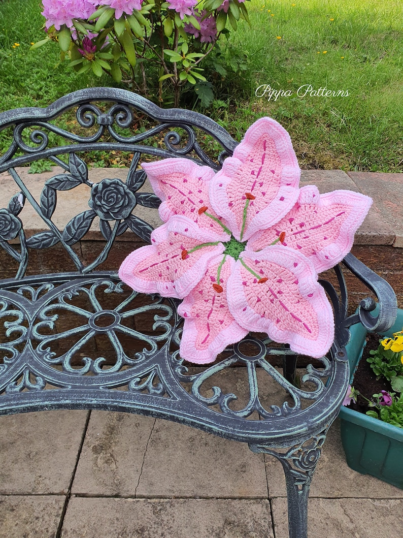 May include: A pink crocheted lily flower cushion resting on a black metal garden bench. The bench is on a stone patio. The text "Pippa Patterns" is visible in the upper right corner of the image.