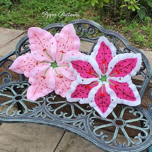 May include: Two crocheted lily flower cushions in pink and white on a black metal bench. The cushions are made with a detailed stitch pattern and have a 3D effect.