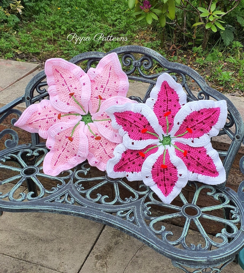 May include: Two crocheted lily flower cushions in pink and white on a black metal bench. The cushions are made with a detailed stitch pattern and have a 3D effect.