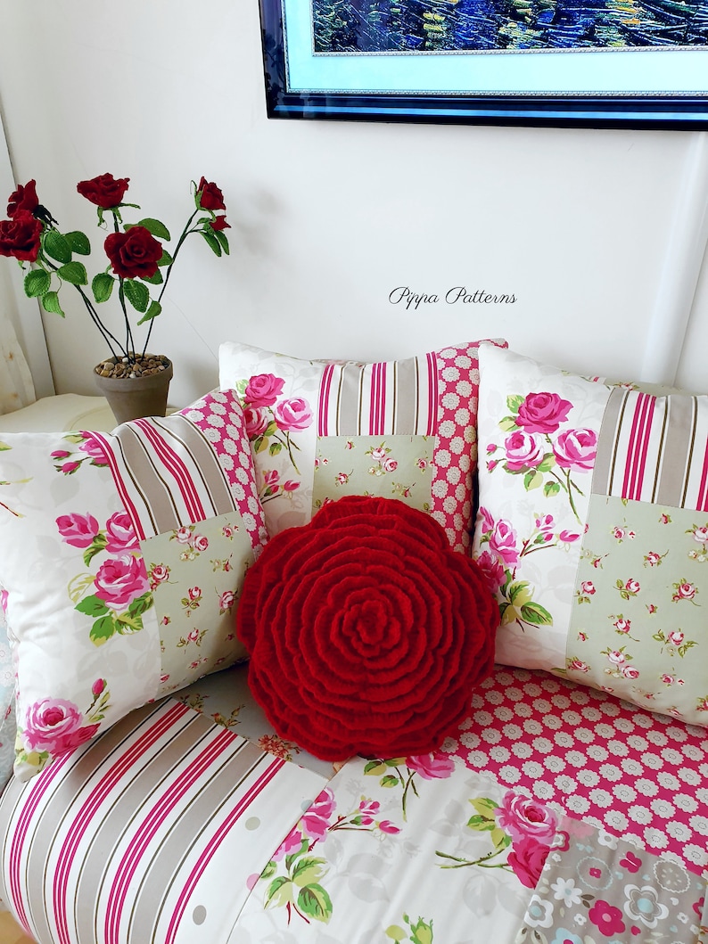 May include: A red, crocheted rose pillow sits on a couch with floral patterned cushions. The couch is covered in a patchwork fabric with pink and white stripes. The text "Pippa Patterns" is visible in the background.