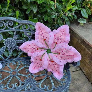 May include: A pink crocheted lily flower cushion with green accents sits on a black metal bench. The cushion is a handmade craft project and features a detailed design.