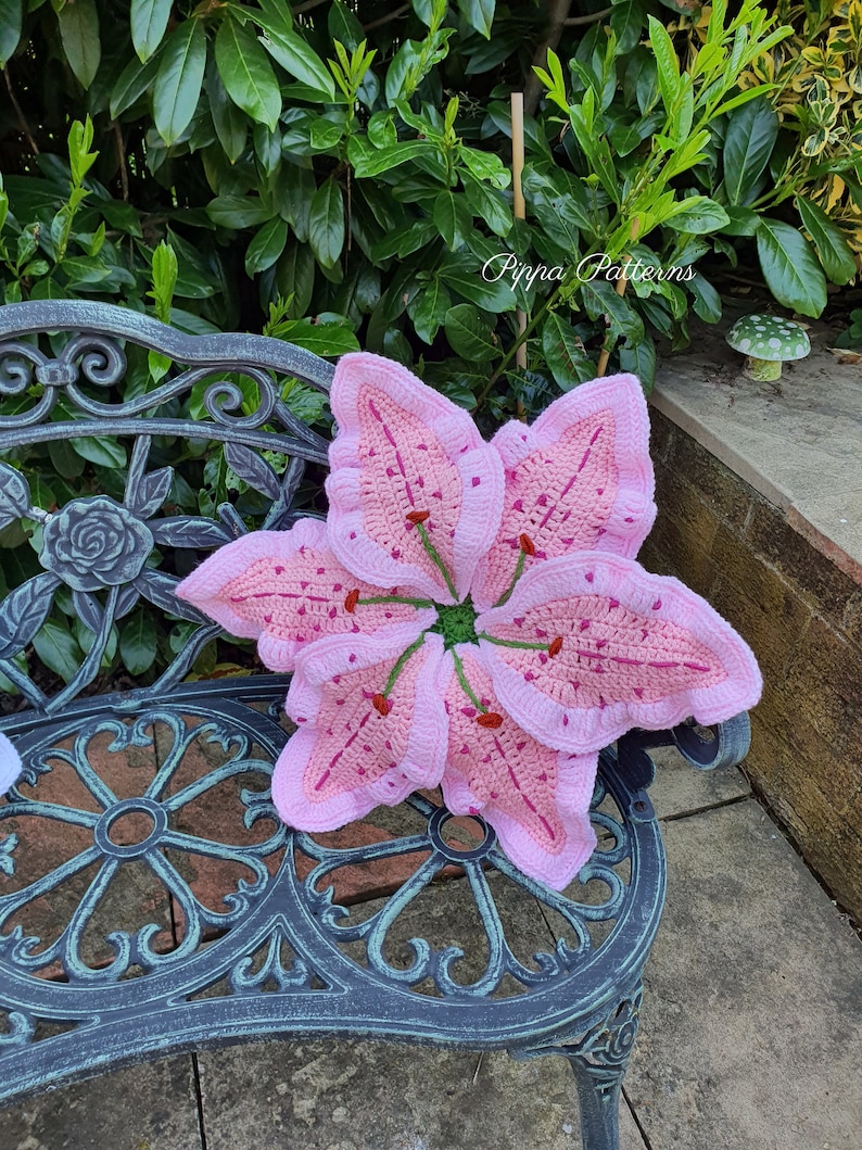 May include: A pink crocheted lily flower cushion with green accents sits on a black metal bench. The cushion is a handmade craft project and features a detailed design.