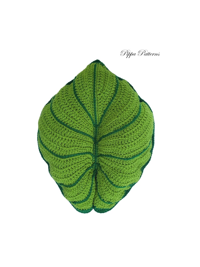May include: A green crocheted leaf pillow with a dark green outline. The pillow is shaped like a large leaf with a central vein and smaller veins branching out.