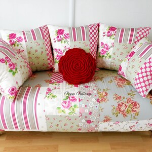 May include: A white and pink patchwork sofa with a red rose-shaped throw pillow. The sofa has a floral pattern and is decorated with several pillows.